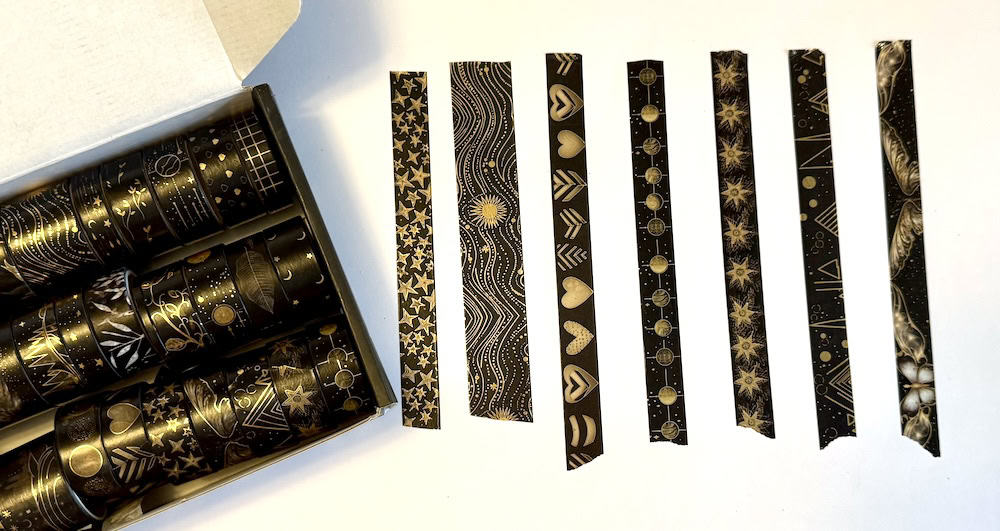 A box with multiple rolls of black tape with gold designs. Nine strips are lined up parallel, featuring various patterns like hearts, stars, and geometric shapes on a white surface.