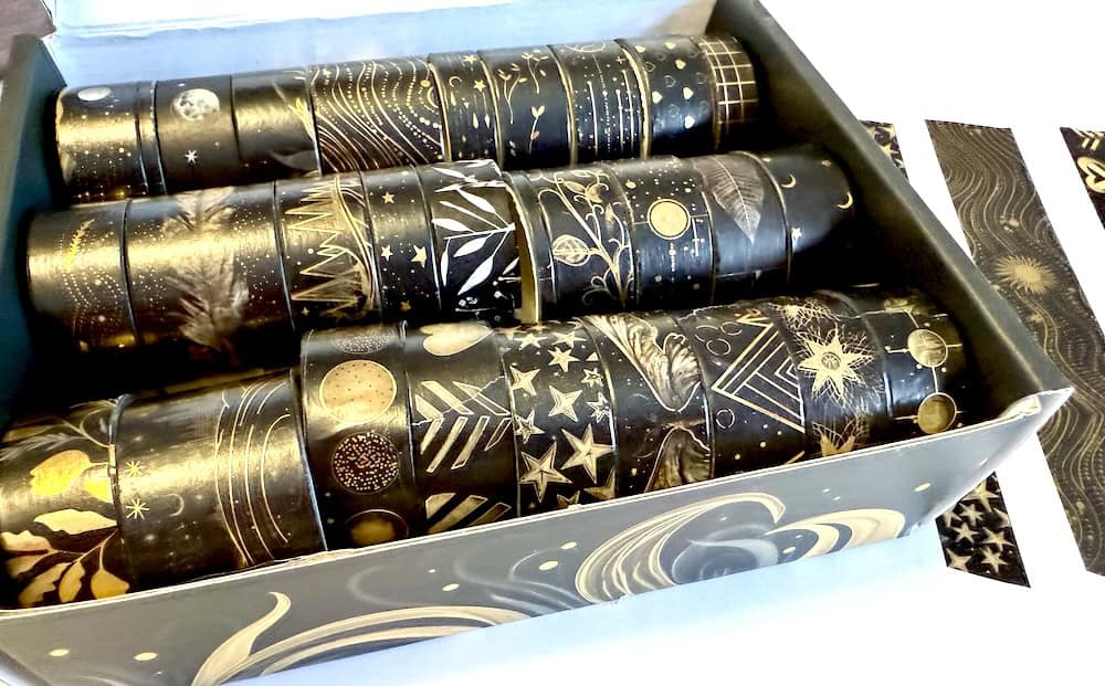 A box contains rolls of black tape with gold designs, featuring celestial and geometric patterns.